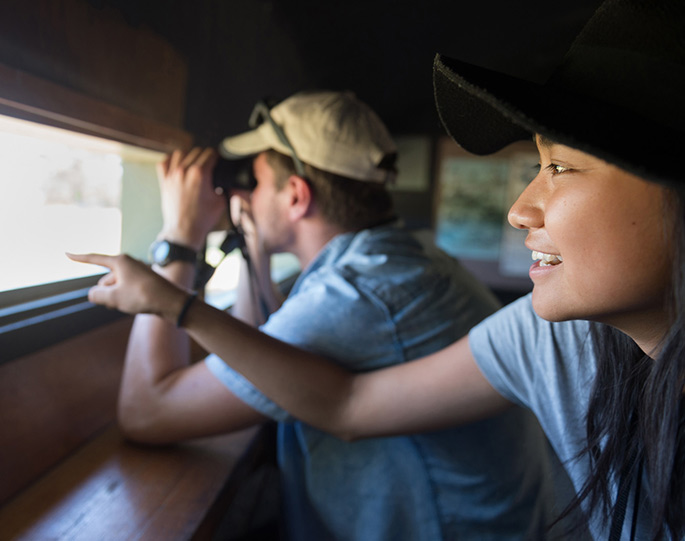 Two students bird watching. One looking through binoculars, while the other points at bird in distance